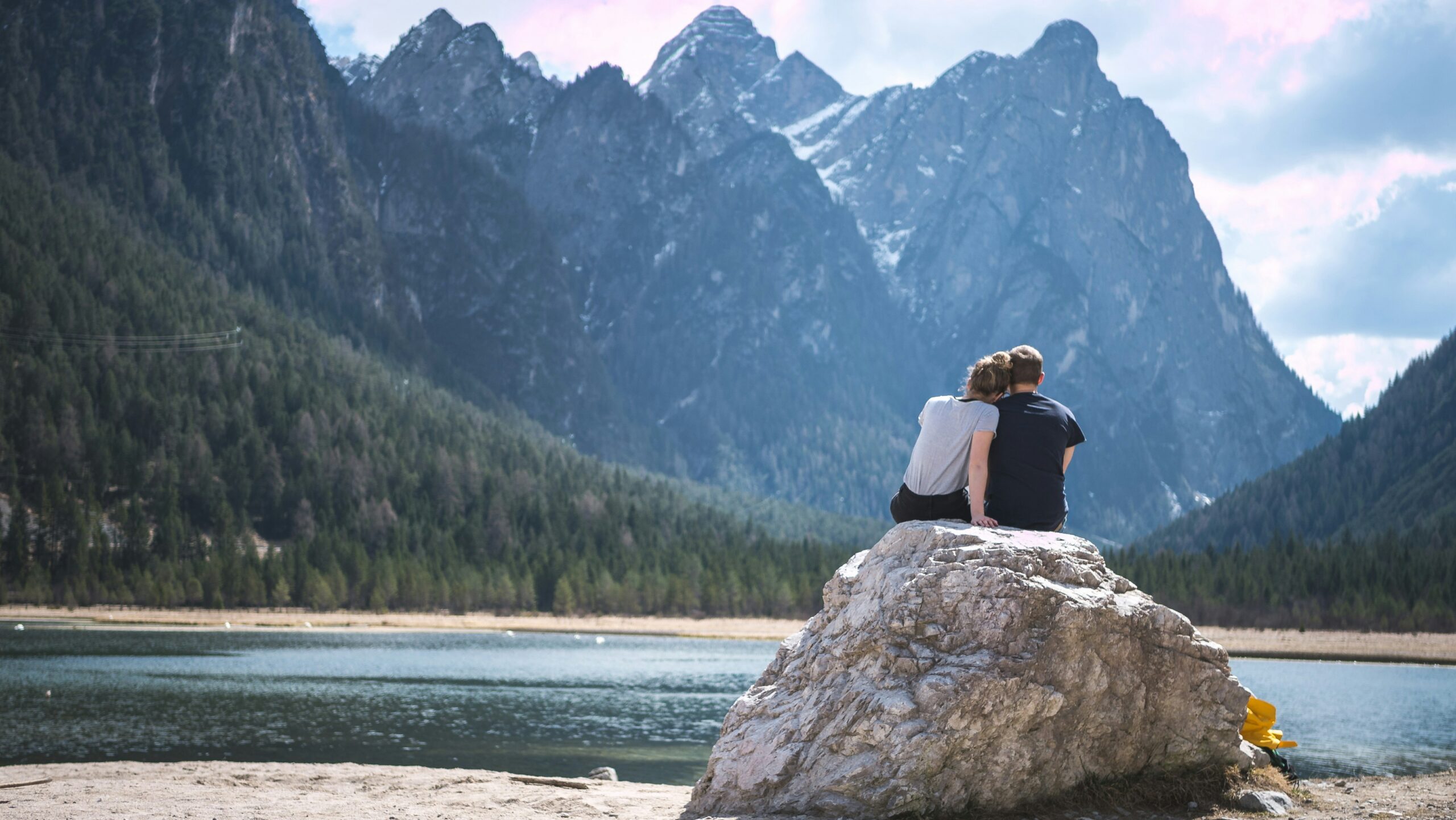 A man and woman sit on a boulder by a lake and mountains.
